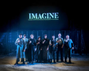 A group of people dressed in poor, raggedy, Victorian-era clothing huddle together and look out towards the audience and camera, signing in BSL as they stand in a dim, cold light. On a screen behind them, the word 'Imagine' almost glows in a cool blue colour.