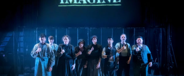 A group of people dressed in poor, raggedy, Victorian-era clothing huddle together and look out towards the audience and camera, signing in BSL as they stand in a dim, cold light. On a screen behind them, the word 'Imagine' almost glows in a cool blue colour.