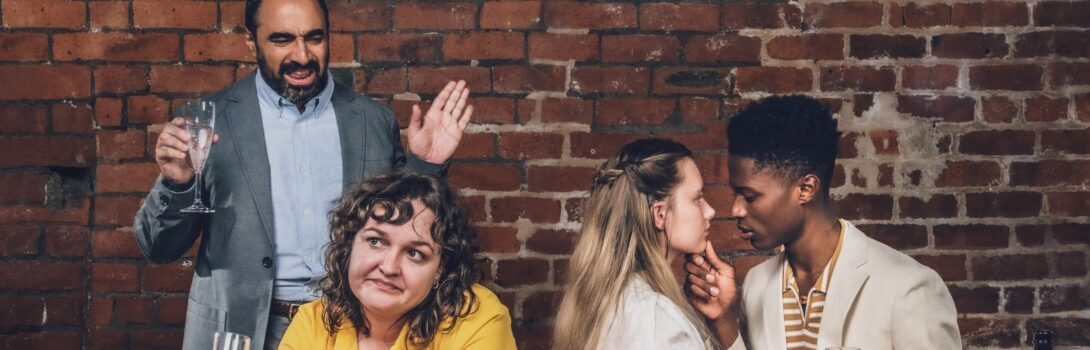 The image shows two men and two women sitting at a long wooden table, their backs against a brick wall. On the left is Benedick, standing up while the others remain seated, and exclaiming something to the back of Beatrice's head, a glass in one hand and his other hand up in defence. Beatrice, wearing a yellow top, faces towards the camera, away from him, tipping her wine glass slightly and looking exasperated. Beside her are Hero and Claudio, engrossed in each other's company, about to kiss.