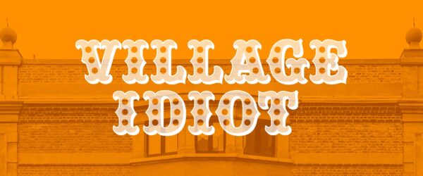 A photo of a grand building, like a town hall, is bathed beneath an orange colour, the words 'Village Idiot' written over the top in an old-fashioned white font.