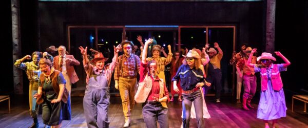 The Company of Much Ado About Nothing. Photo by Johan Persson. The full company of performers are line dancing and are all dressed in Country and Western clothing. There are colourful lights and people lift their arms in the air as they dance.