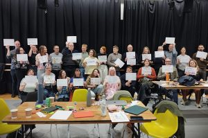 The first cohort of the Ramps on the Moon cohort - about 25 people - are all gathered as a crowd in front of a floor to ceiling black curtain. They are all holding a placard in a silent protest against ableism.
