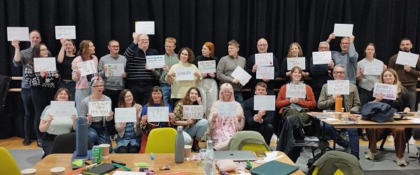 The first cohort of the Ramps on the Moon cohort - about 25 people - are all gathered as a crowd in front of a floor to ceiling black curtain. They are all holding a placard in a silent protest against ableism.
