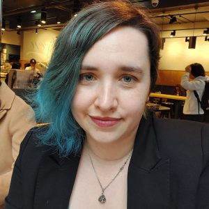 A head shot of Miranda, she has a blue bob and is wearing a black jacket and a pendant round her neck