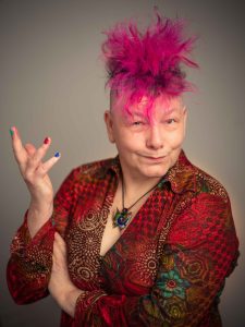 Caronline Parker is smiling into the lens, she has fabulous pink spkey short hair and is wearing a deep red patterned shirt - her right arm is raises in a questioning way