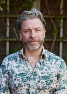 David Belwood head and shoulders pic. David is looking into the lens of the camera, he has a half smile and half serious expression on his face. He has a beard and is wearing a green leaf pattern shirt
