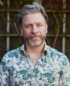 David Belwood head and shoulders pic. David is looking into the lens of the camera, he has a half smile and half serious expression on his face. He has a beard and is wearing a green leaf pattern shirt