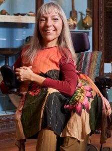 Esther Fox is sitting in her chair, smiling at the camera - she has long blonde hair and is waring red and orange dress