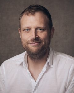 A head and shoulders photo of Tom Bird, He is smiing at the camera. He has short light brown hair and a short beard. He is wearing a white shirt