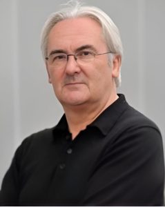 A photo of Stuart Rogers. A head and shoulders shot - Stuart has short white/grey hair, he is wearing thin rimmed spectacles. He is wearing a black polo tshirt
