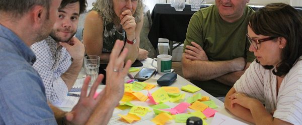 A group of the cohort sit around a table in deep discussion, with lots of post-it notes in front of them. They are working on The Social Model of Disability