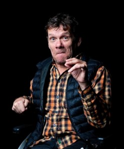 Jamie has his left arm raised and has a smile on his face. He has dark brown wavy short hair, and is wearing a checked shirt and a puffa jacket