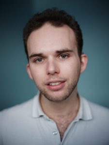 Head and shoulder image of Max Fairley, he has short brown wavy hair and is wearing a light great collared shirt