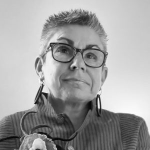 Black and white image of Michele Taylor. She has short styled chopped hair, and is wearning black rimmed glasses. She is looking slightly upwards with an interested smile
